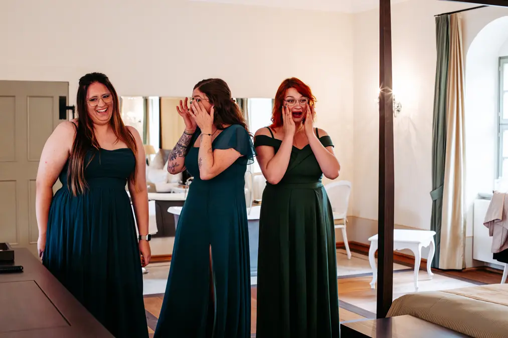 Drei Frauen in passenden blaugrünen Kleidern stehen in einem Raum und reagieren überrascht und erfreut. Zwei halten sich den Mund zu, während die dritte breit lächelt. Der Raum hat einen Holzboden, weiße Wände und große Fenster.