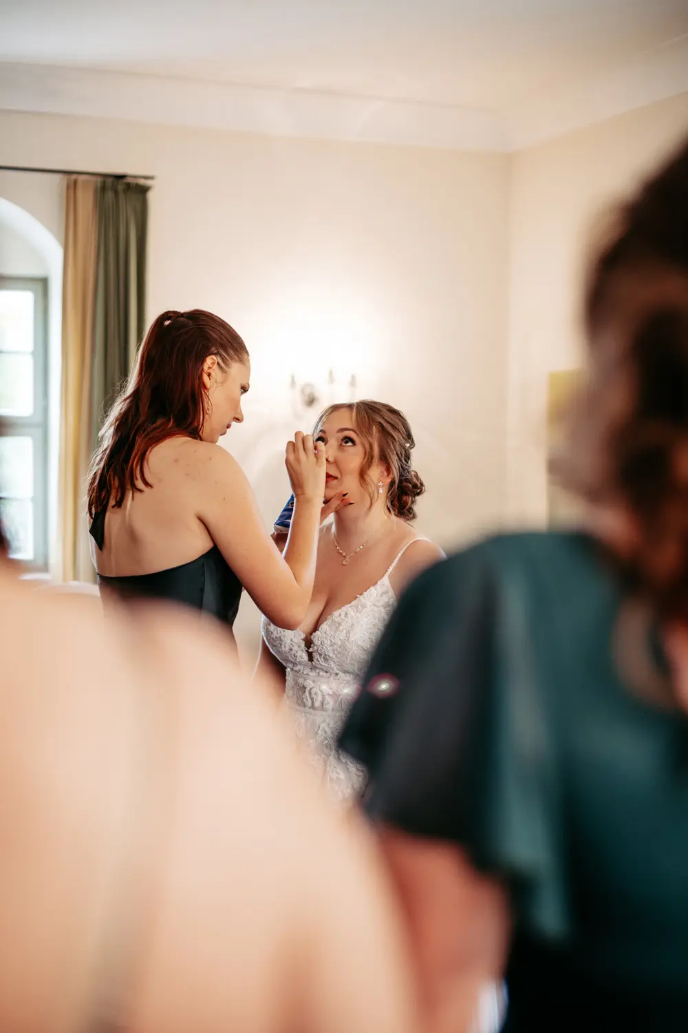 Eine Frau in einem schwarzen Kleid hilft einer Braut in einem weißen Kleid beim Schminken und berührt dabei sanft ihr Gesicht in einem sanft beleuchteten Raum mit einem Fenster und hellen Wänden. Andere Personen sind unscharf im Vordergrund zu sehen.