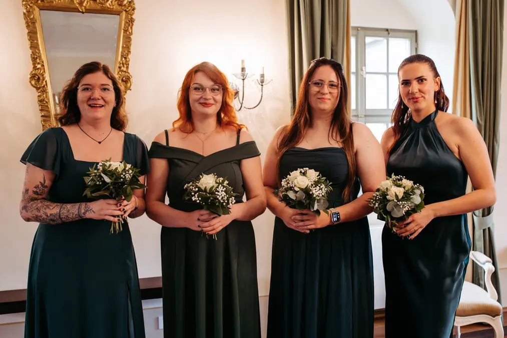 Vier Frauen in dunkelgrünen Kleidern stehen in einem Innenraum nebeneinander und halten kleine Sträuße aus weißen Blumen und Grünzeug. Sie lächeln, im Hintergrund sind ein Spiegel, ein Wandleuchter und ein Fenster zu sehen.