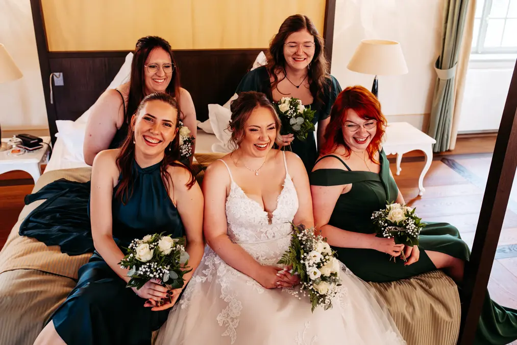 Eine Braut in einem weißen Kleid sitzt auf einem Bett, umgeben von vier Brautjungfern in dunkelgrünen Kleidern. Alle lächeln und halten kleine Sträuße aus weißen Blumen in einem warm beleuchteten Raum.