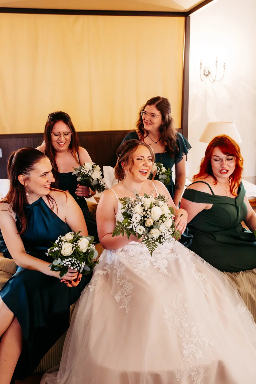 Eine Braut in einem weißen Kleid sitzt lachend auf einem Bett und hält einen Blumenstrauß in der Hand, umgeben von vier Brautjungfern in grünen Kleidern, die alle lächeln und kleine Blumenarrangements halten.