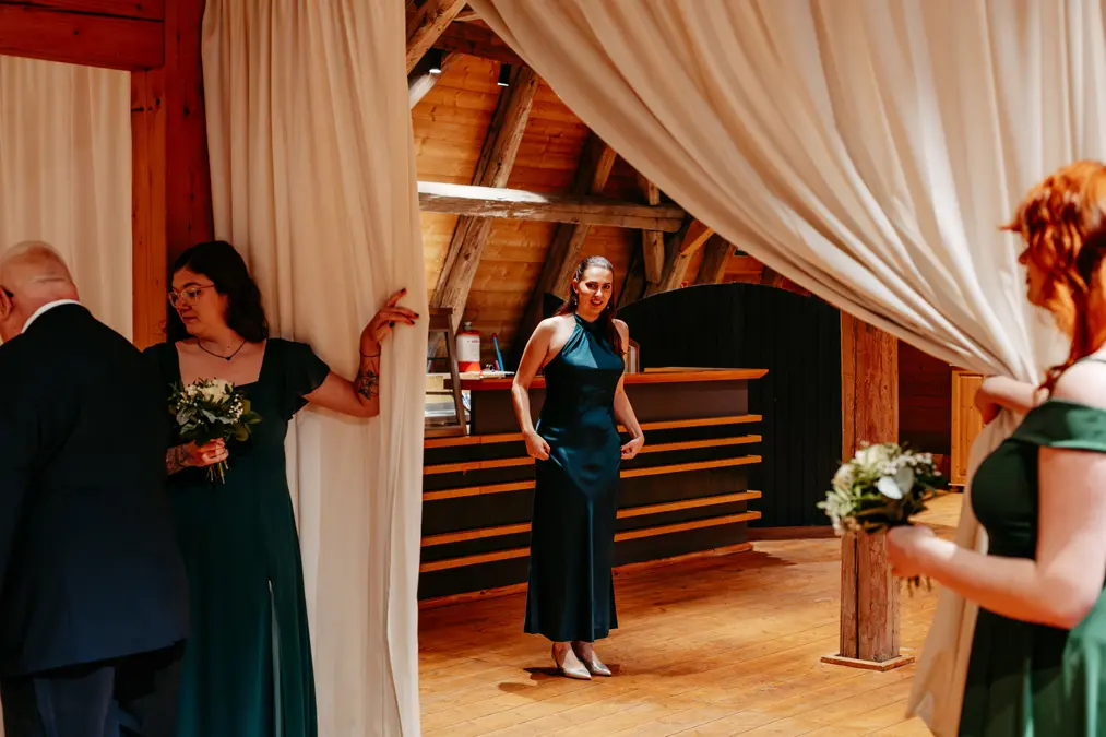 Drei Frauen in dunkelgrünen Kleidern und ein Mann stehen in einem hölzernen Saal. Zwei Frauen halten Blumensträuße und teilen weiße Vorhänge, während eine andere Frau lächelnd im Hintergrund steht. Warmes Sonnenlicht strömt von oben herein.