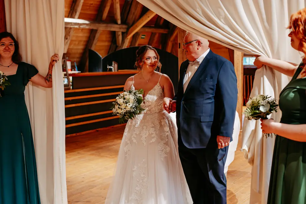 Eine Braut in einem weißen Spitzenkleid mit einem Blumenstrauß in der Hand geht Arm in Arm mit einem älteren Mann in einem marineblauen Anzug. Zwei Frauen in grünen Kleidern halten weiße Vorhänge offen, als sie einen warm beleuchteten Holzsaal betreten.
