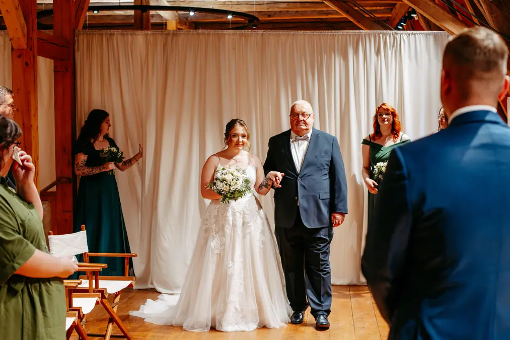 Eine Braut in einem weißen Kleid und mit einem Blumenstrauß in der Hand geht Arm in Arm mit einem älteren Mann im Anzug den Gang entlang, während die Hochzeitsgäste und Brautjungfern in grünen Kleidern zusehen.