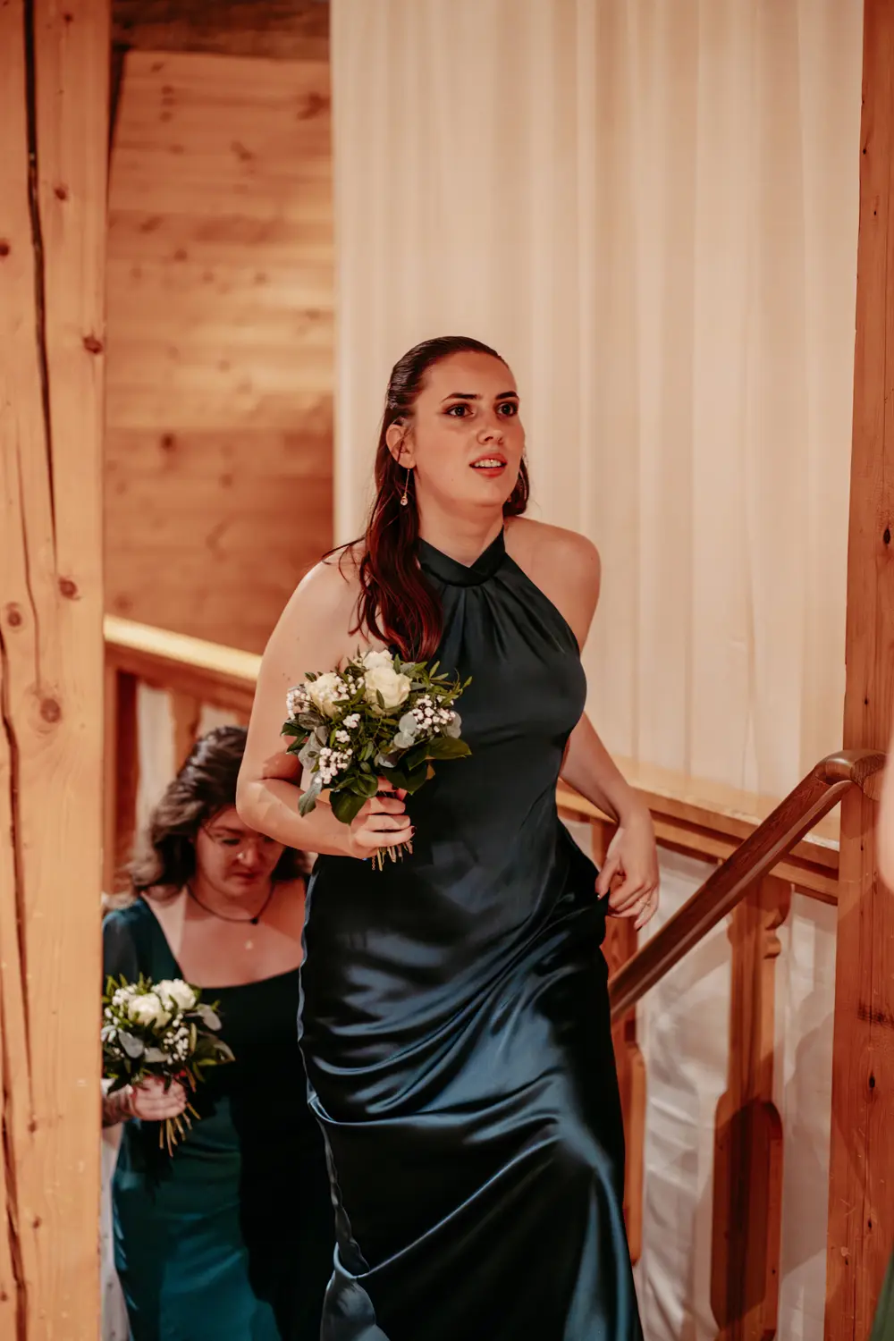 Zwei Frauen in dunkelgrünen Kleidern halten kleine Blumensträuße in der Hand und gehen eine Holztreppe hinauf, möglicherweise auf einer Hochzeit. Die vordere Frau blickt mit konzentriertem Blick nach vorn. Die warme Beleuchtung hebt die hölzerne Umgebung hervor.
