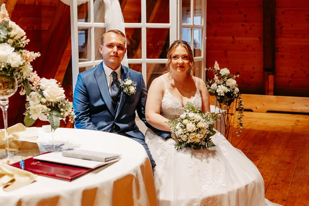 Eine Braut in einem weißen Kleid und ein Bräutigam in einem blauen Anzug sitzen Seite an Seite in einem Raum, halten Sträuße und lächeln, umgeben von weißen Blumen und Holzdekor.