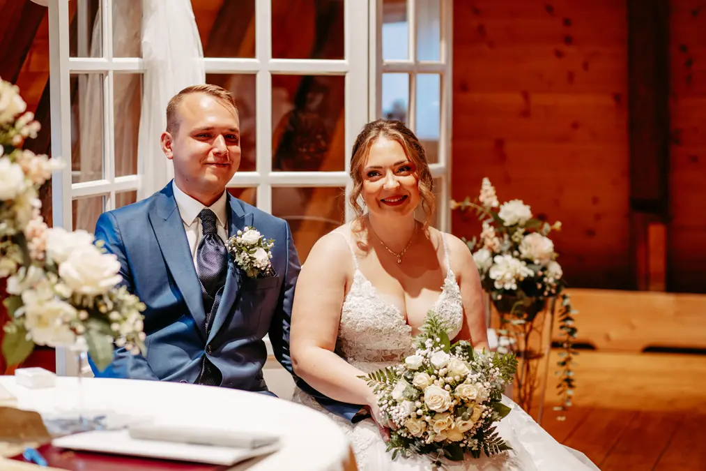 Eine Braut in einem weißen Kleid und ein Bräutigam in einem blauen Anzug sitzen lächelnd nebeneinander und halten einen Strauß weißer Blumen, umgeben von Blumenarrangements in einem warm beleuchteten, hölzernen Raum.