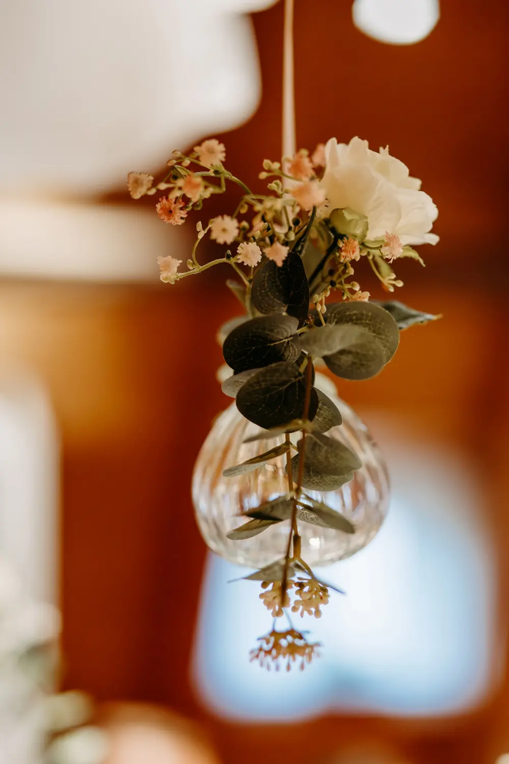 Eine gläserne, zwiebelförmige Vase hängt kopfüber mit kleinen rosa und weißen Blüten und grünen Blättern vor einem warmen, verschwommenen Hintergrund.