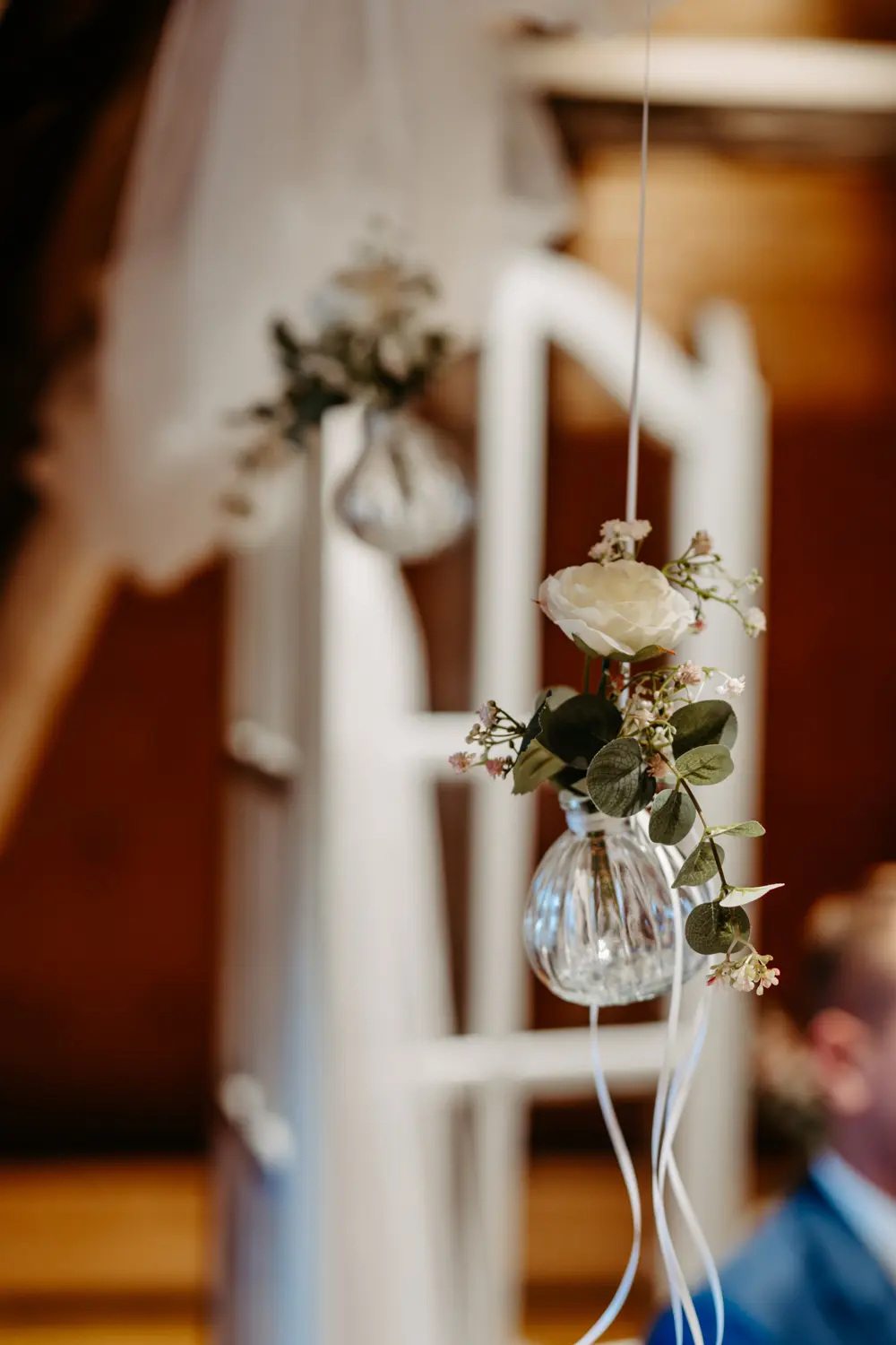 Eine Nahaufnahme einer kleinen Glasvase, die an einem weißen Band aufgehängt ist und eine weiße Rose und Grünzeug enthält, mit einem unscharfen Hintergrund, der eine weitere ähnliche Vase und eine weiche, warme Beleuchtung zeigt.