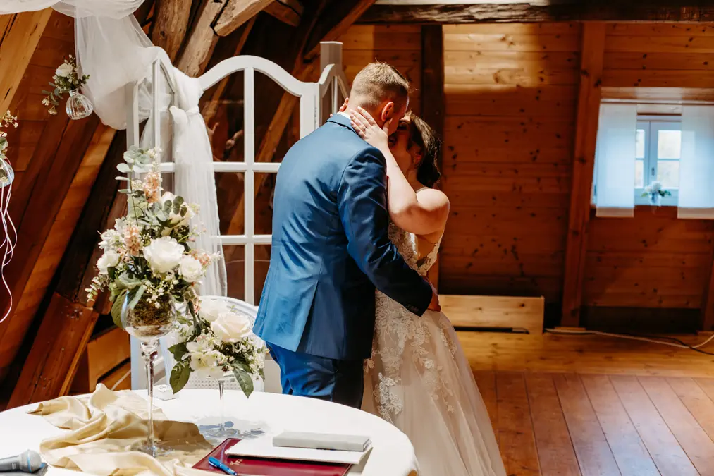 Eine Braut und ein Bräutigam küssen sich in einem rustikalen, mit Blumen und weißen Vorhängen geschmückten Holzzimmer und stehen neben einem kleinen Tisch mit einem Gästebuch und einem Blumengesteck. Licht strömt durch ein Fenster herein.