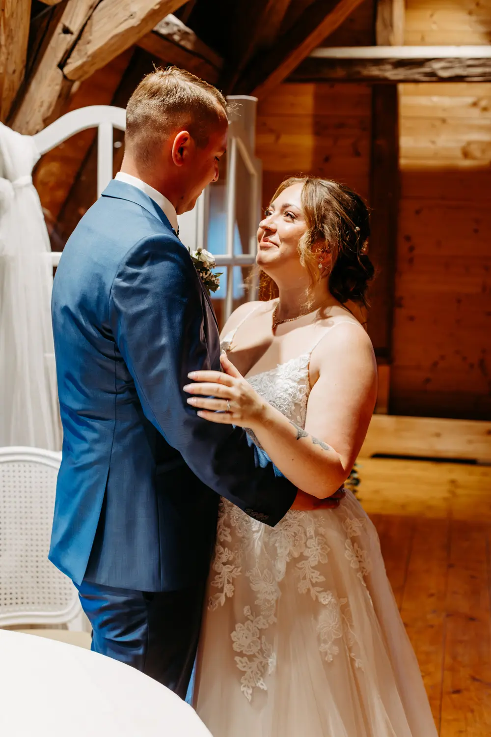 Eine Braut im Spitzenhochzeitskleid und ein Bräutigam im blauen Anzug werfen sich einen liebevollen Blick zu, während sie in einem warm beleuchteten, rustikalen Holzlokal miteinander tanzen.