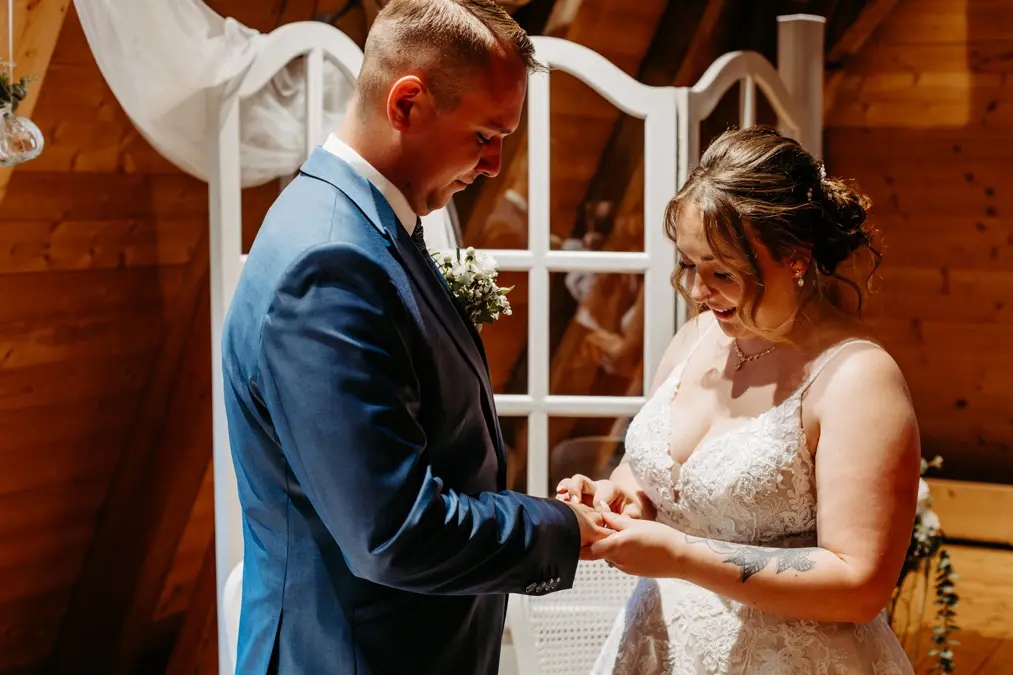 Eine Braut und ein Bräutigam stehen eng beieinander und lächeln, während die Braut dem Bräutigam während ihrer Hochzeitszeremonie einen Ring an den Finger steckt. Die Umgebung zeichnet sich durch warme Holzwände und sanftes natürliches Licht aus.