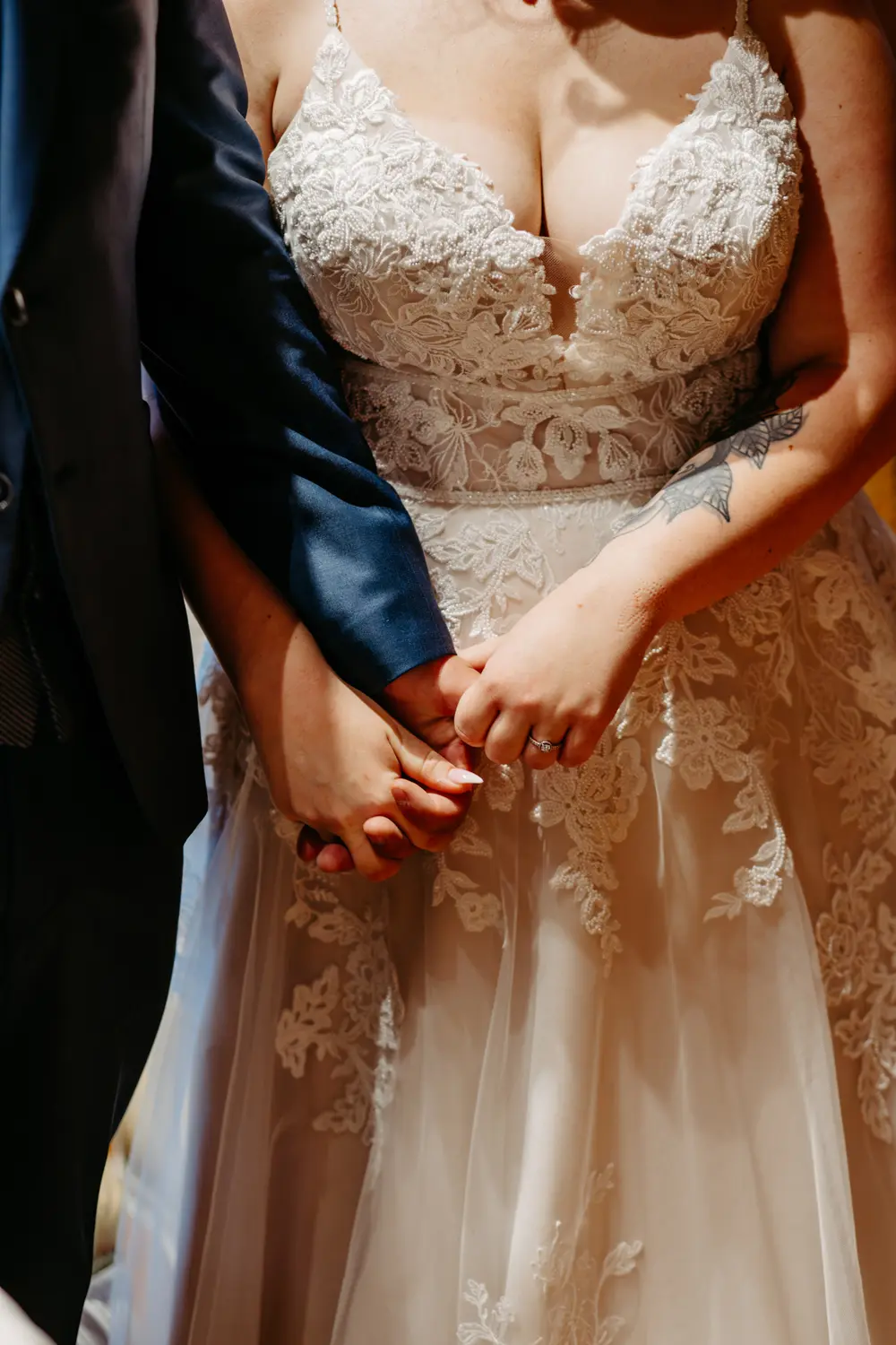 Nahaufnahme einer Braut und eines Bräutigams, die sich an den Händen halten. Die Braut trägt ein Hochzeitskleid aus Spitze mit Blumenmuster, der Bräutigam einen dunklen Anzug. Das Tattoo der Braut ist auf ihrem Arm zu sehen.
