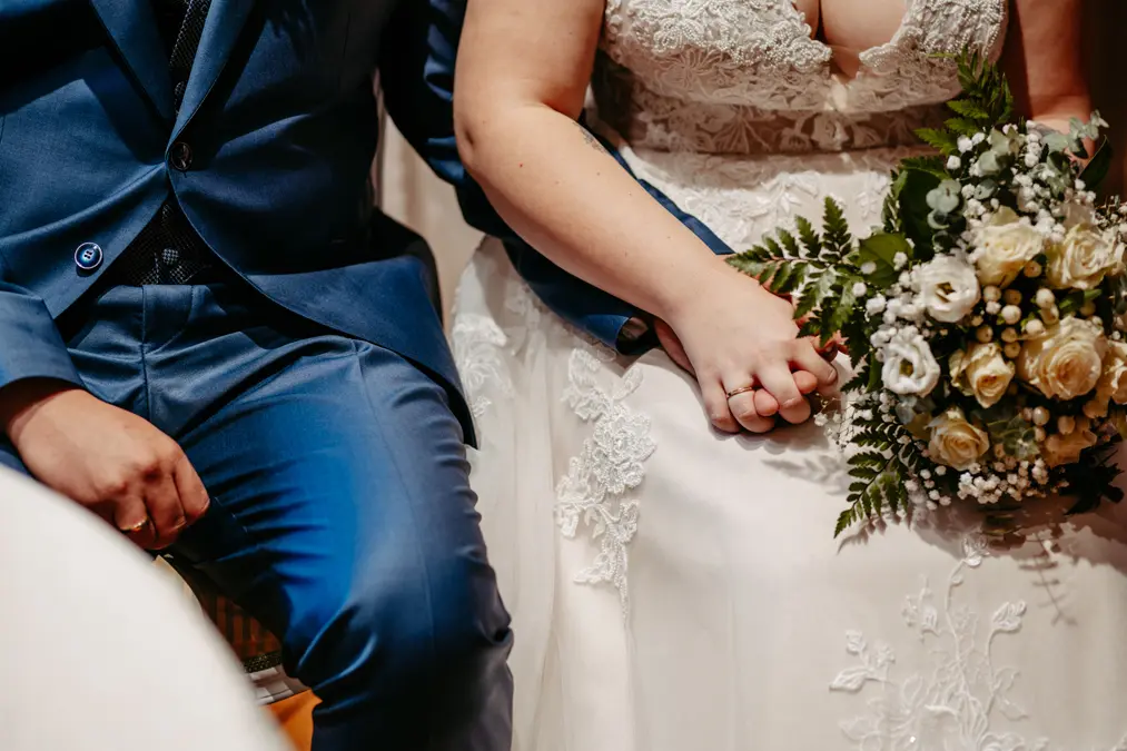 Nahaufnahme von Braut und Bräutigam, die nebeneinander sitzen und sich an den Händen halten. Die Braut trägt ein weißes Spitzenkleid und hält einen Strauß weißer Blumen; der Bräutigam trägt einen blauen Anzug.