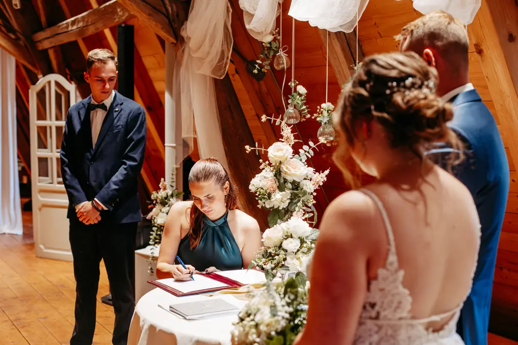 Eine Frau in einem blaugrünen Kleid unterschreibt ein Dokument an einem runden, blumengeschmückten Tisch, während eine Braut und ein Bräutigam daneben stehen. Ein weiterer Mann im Anzug steht in einem warm beleuchteten, rustikalen Holzzimmer an der Seite.