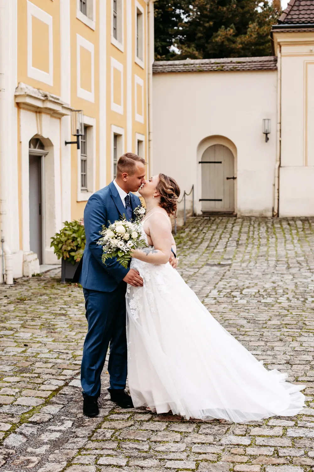 Eine Braut in einem weißen Kleid und ein Bräutigam in einem blauen Anzug küssen sich vor einem gelben und weißen Gebäude mit gewölbten Fenstern und Türen in einem Hof mit Kopfsteinpflaster. Die Braut hält einen Strauß mit weißen Blumen.