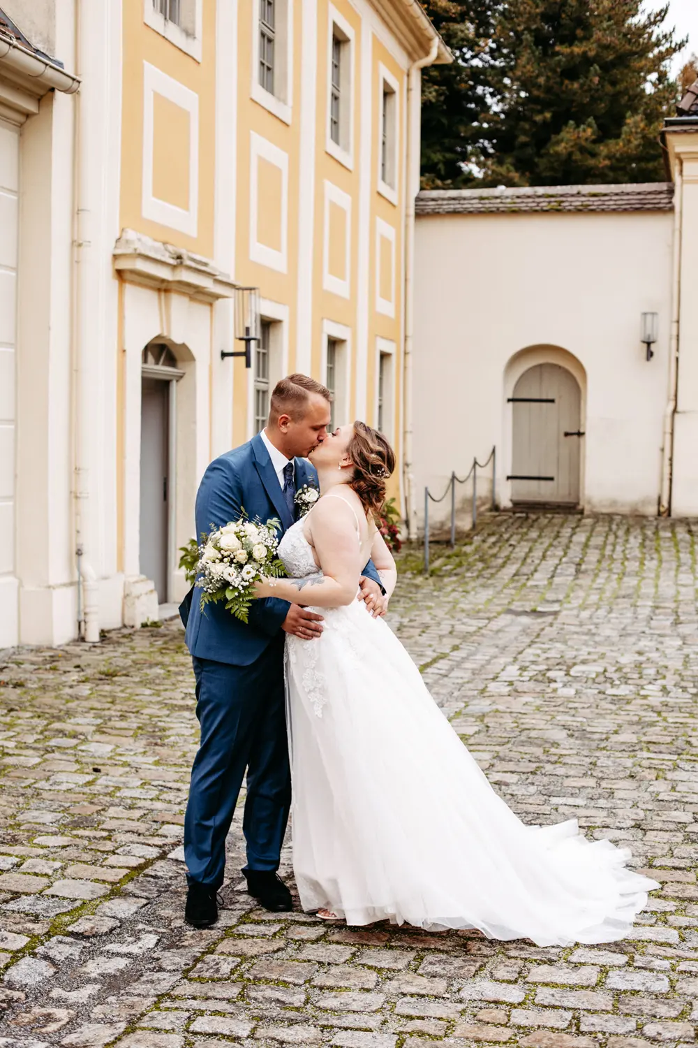 Eine Braut in einem weißen Kleid und ein Bräutigam in einem blauen Anzug umarmen sich vor einem gelb-weißen Gebäude mit Kopfsteinpflaster.