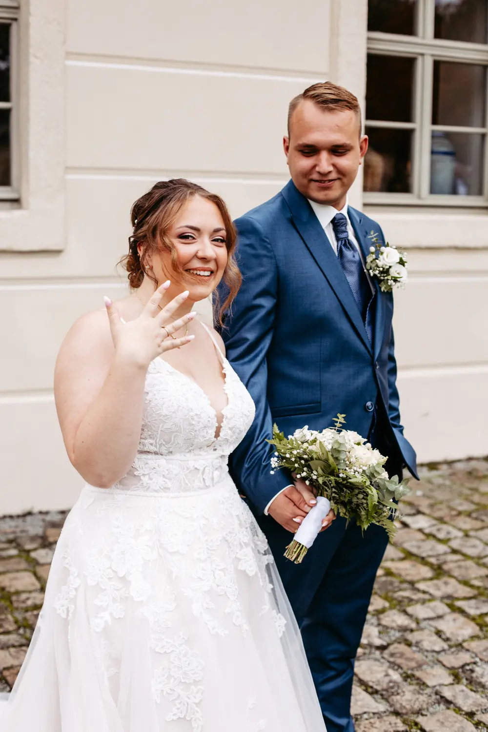 Eine lächelnde Braut in einem weißen Spitzenkleid zeigt ihren Ring, während sie die Hand des Bräutigams in einem blauen Anzug hält. Beide stehen auf einem kopfsteingepflasterten Weg vor einem cremefarbenen Gebäude mit Fenstern.