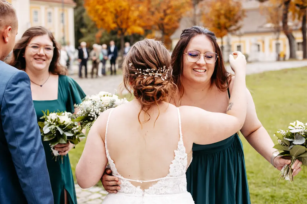 Eine Braut in einem weißen Spitzenkleid umarmt eine Brautjungfer in einem grünen Kleid im Freien. Eine andere Brautjungfer steht in der Nähe und hält Blumen. Im unscharfen Hintergrund sind Menschen und Herbstbäume zu sehen.
