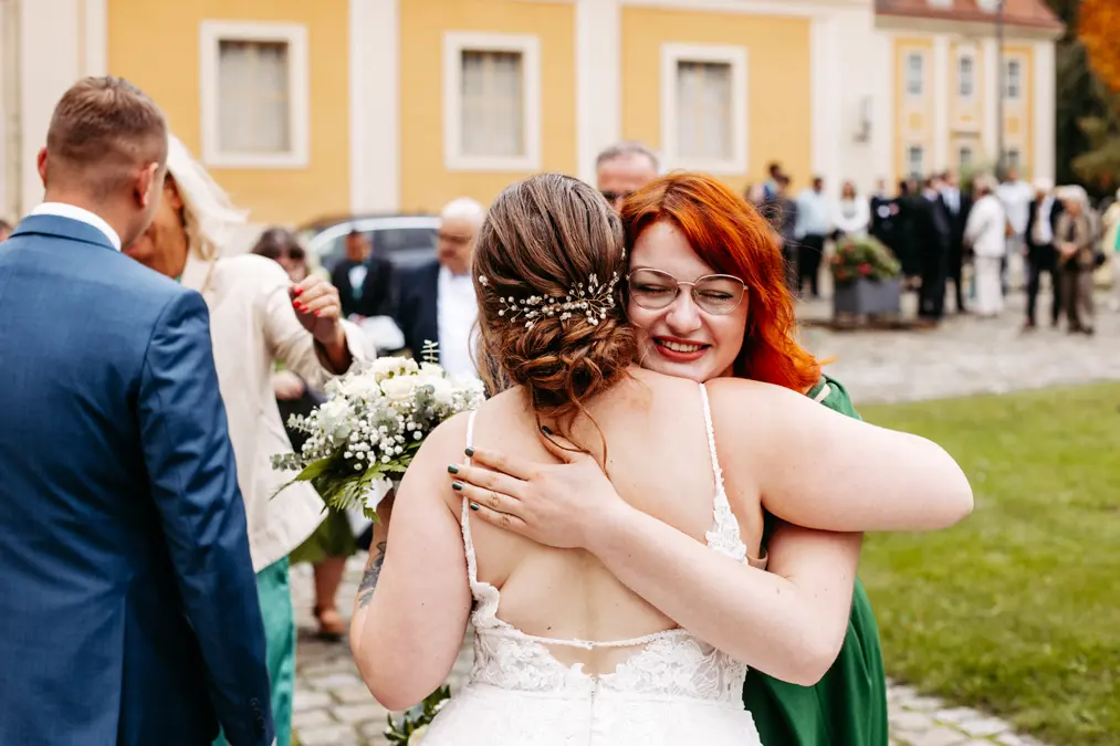 Eine Braut in einem weißen Kleid mit geblümtem Haarschmuck umarmt eine lächelnde Frau mit roten Haaren und Brille während einer Hochzeitsfeier im Freien, mit Gästen und einem gelben Gebäude im Hintergrund.
