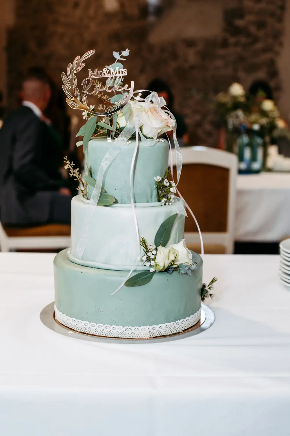 Eine dreistöckige Hochzeitstorte mit grünem und weißem Zuckerguss, verziert mit weißen Blumen, Grünzeug, Bändern und einem Mr & Mrs Topper auf einem weißen Tischtuch.