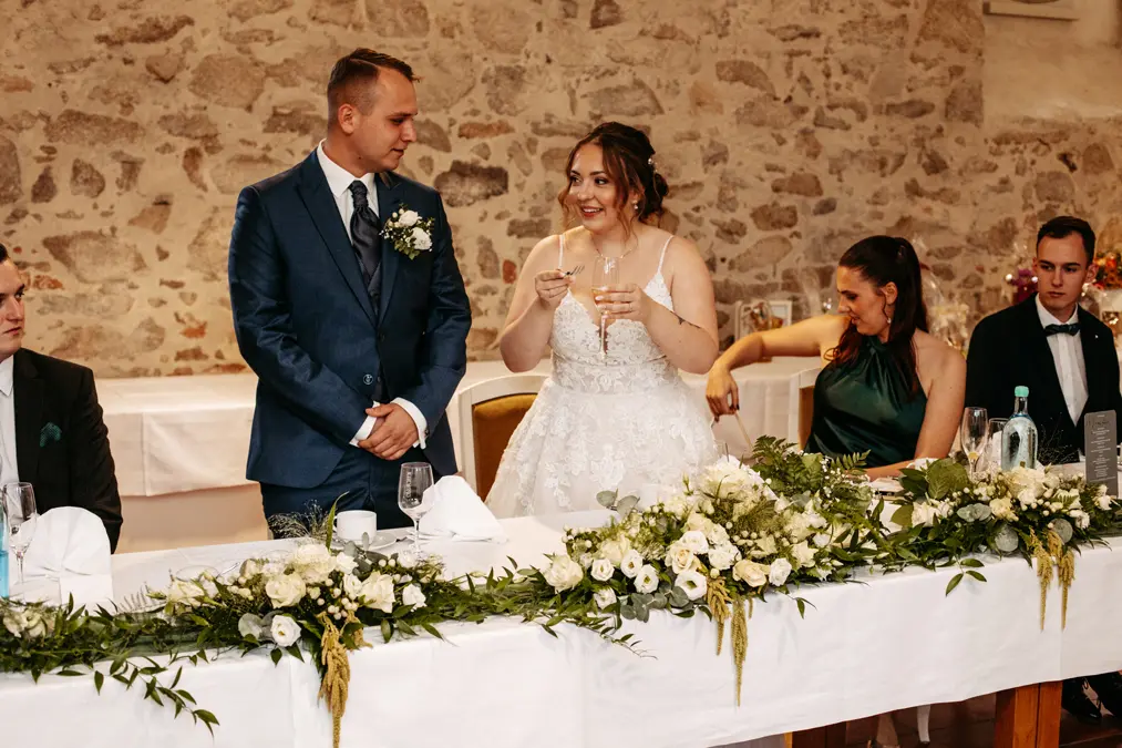 Braut und Bräutigam stehen hinter einem geschmückten Tisch und lächeln, während die Braut zwei Gläser hochhält. Brautjungfern und Trauzeugen sitzen in der Nähe, mit Blumenarrangements und Grünzeug vor ihnen, vor dem Hintergrund einer Steinmauer.