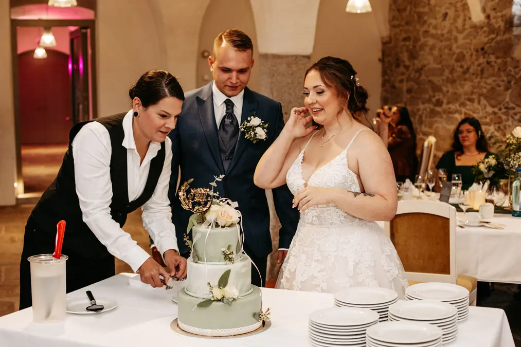 Braut und Bräutigam stehen neben einer Frau, die ihre Hochzeitstorte bei einem Empfang anschneidet. Die Braut trägt ein weißes Kleid, der Bräutigam einen blauen Anzug, und auf dem Tisch neben der Torte liegt ein Stapel von Tellern. Die Gäste sitzen im Hintergrund.