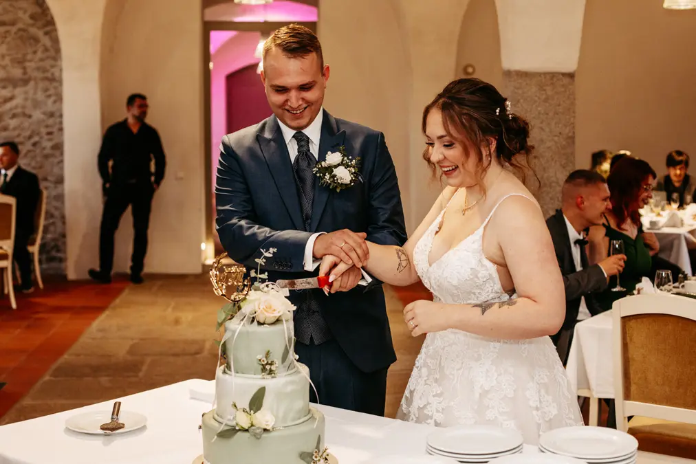 Eine lächelnde Braut und ein lächelnder Bräutigam schneiden bei ihrem Empfang gemeinsam eine mehrstöckige Hochzeitstorte an. Die Braut trägt ein weißes Kleid und der Bräutigam einen blauen Anzug. Die Gäste sitzen an Tischen im Hintergrund.