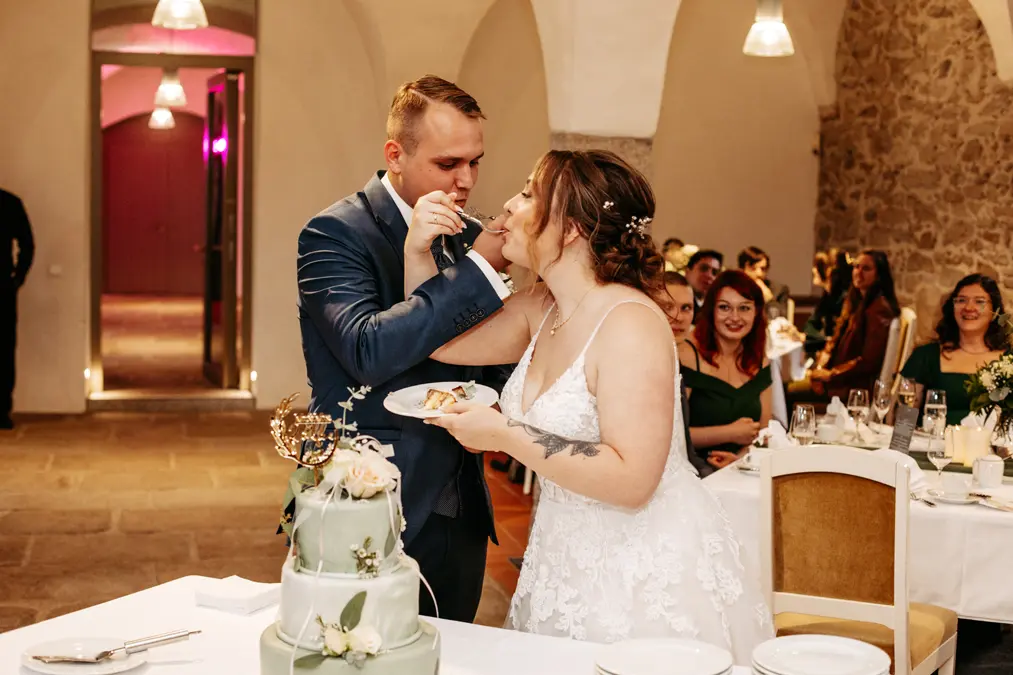 Braut und Bräutigam stehen bei ihrem Hochzeitsempfang beieinander, während der Bräutigam der Braut vorsichtig die Torte reicht. Die an Tischen sitzenden Gäste schauen zu und lächeln in dem warm beleuchteten, von Steinmauern umgebenen Lokal.