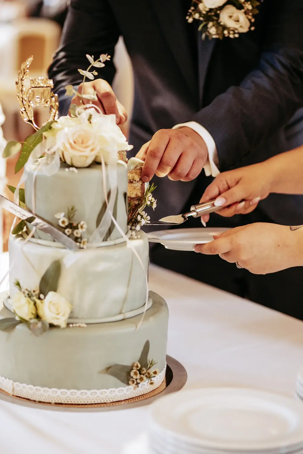 Ein Paar schneidet ein Stück von einer dreistöckigen Hochzeitstorte mit weißem und grünem Fondant, frischen weißen Rosen und Grünzeug ab und legt das Stück gemeinsam auf einen Teller.