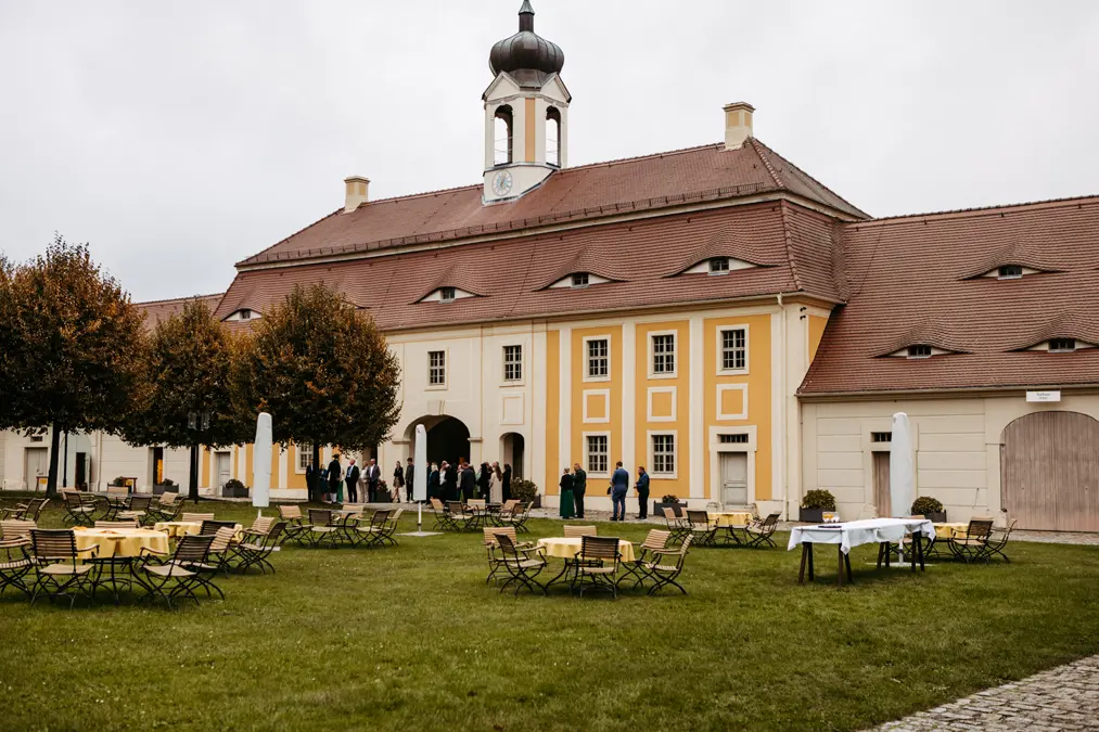 Ein großes historisches Gebäude mit gelben und weißen Wänden und einem Uhrenturm steht hinter einer offenen Rasenfläche mit leeren Tischen und Stühlen. In der Nähe des Eingangs haben sich bei bewölktem Himmel Menschen versammelt.