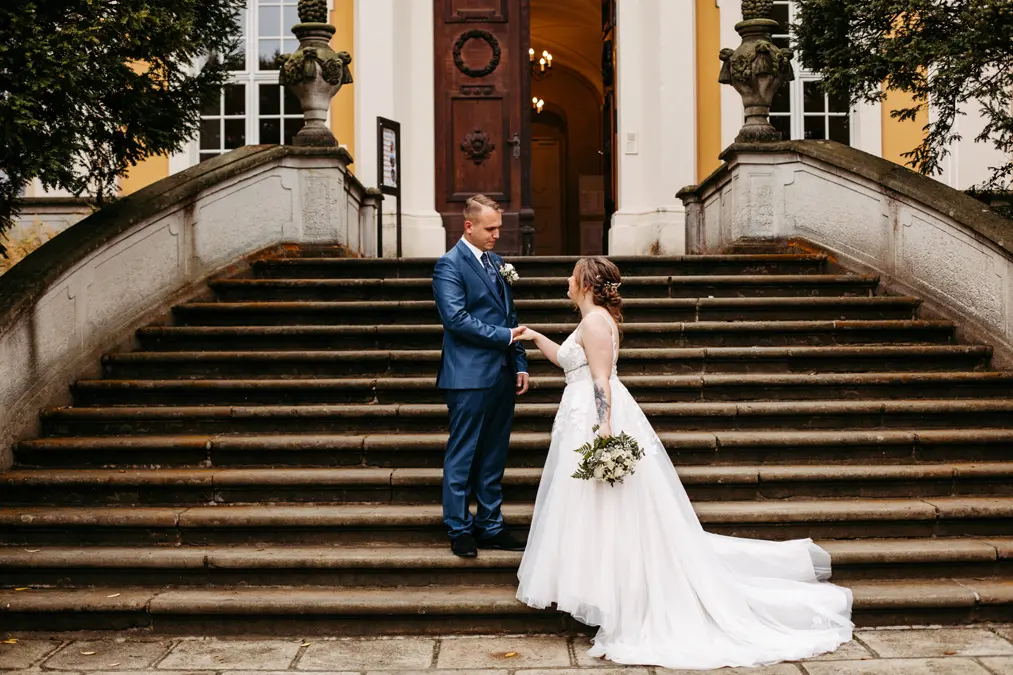 Eine Braut in einem weißen Kleid und ein Bräutigam in einem blauen Anzug stehen sich händchenhaltend auf einer Steintreppe im Freien gegenüber, mit einem großen Gebäudeeingang hinter ihnen. Die Braut hält einen Blumenstrauß in der Hand.