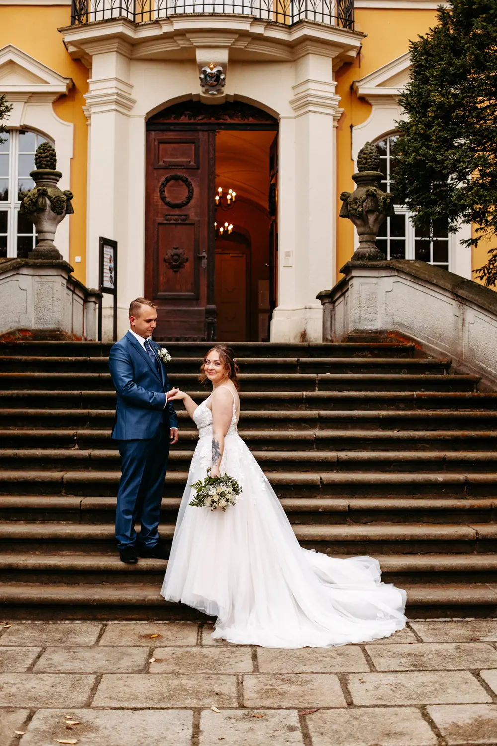 Eine Braut in einem weißen Kleid mit einem Blumenstrauß in der Hand steht mit ihrem Bräutigam in einem blauen Anzug auf Steinstufen vor einem großen Gebäude mit einer Holztür und gelben Wänden.