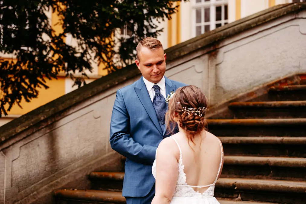 Ein Bräutigam in einem blauen Anzug lächelt eine Braut in einem weißen Kleid mit offenem Rücken und dekorativen Haarnadeln an, als sie gemeinsam im Freien vor einer Steintreppe stehen.