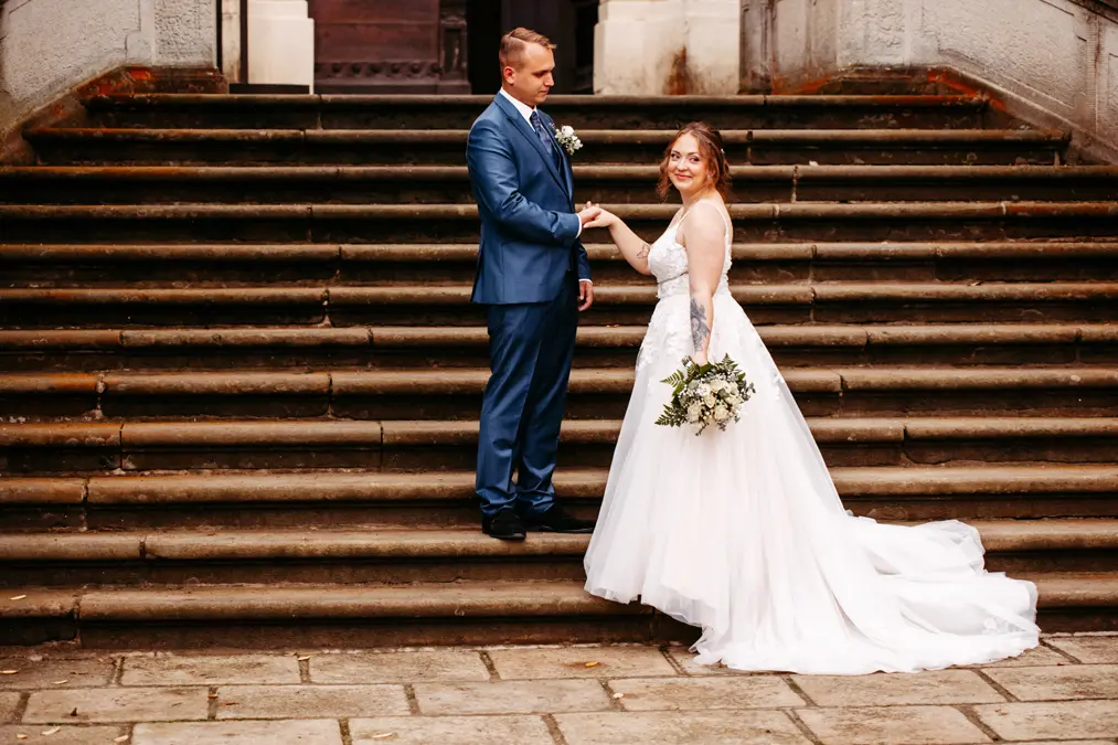 Eine Braut in einem weißen Kleid hält einen Blumenstrauß in der Hand und lächelt, während sie auf steinernen Stufen steht und sich mit ihrem Bräutigam in einem blauen Anzug an den Händen hält. Sie stehen sich vor einem historischen Gebäude gegenüber.