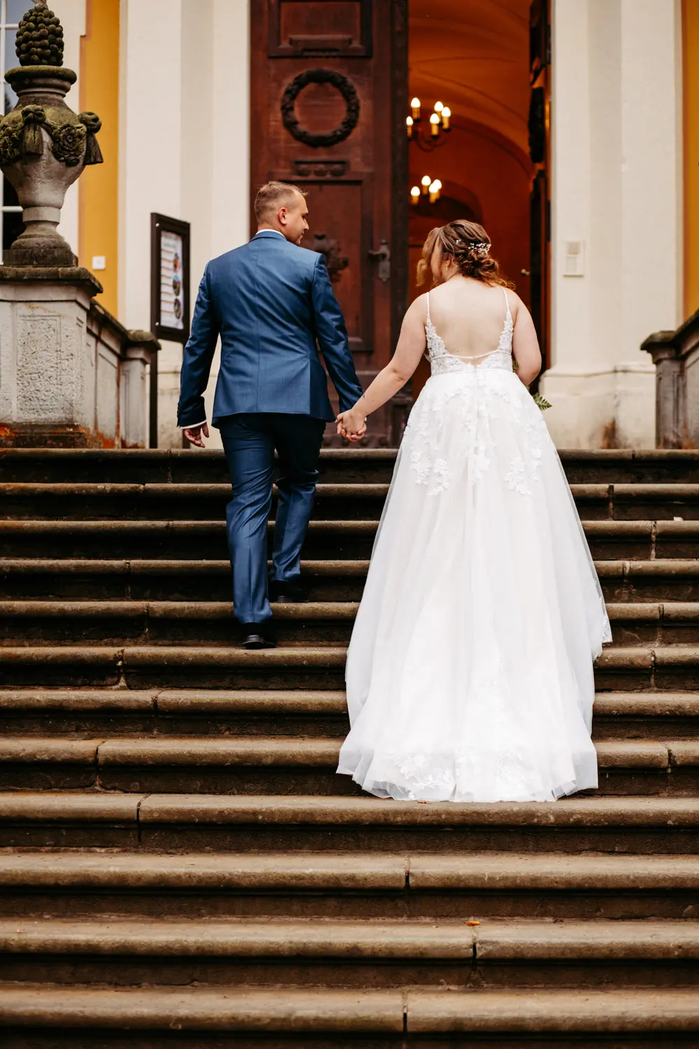 Eine Braut in einem weißen Kleid und ein Bräutigam in einem blauen Anzug halten sich an den Händen, während sie über Steinstufen auf einen Gebäudeeingang zugehen, von hinten gesehen.