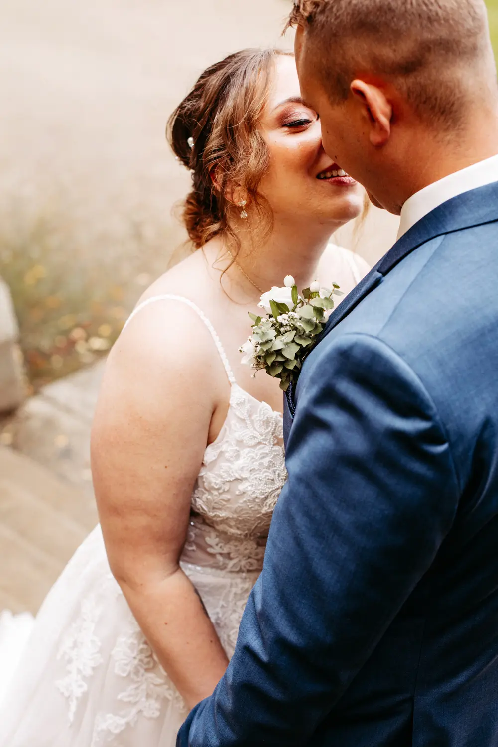 Eine Braut in einem weißen Spitzenkleid und ein Bräutigam in einem blauen Anzug stehen dicht beieinander, lächeln und küssen sich an ihrem Hochzeitstag im Freien.