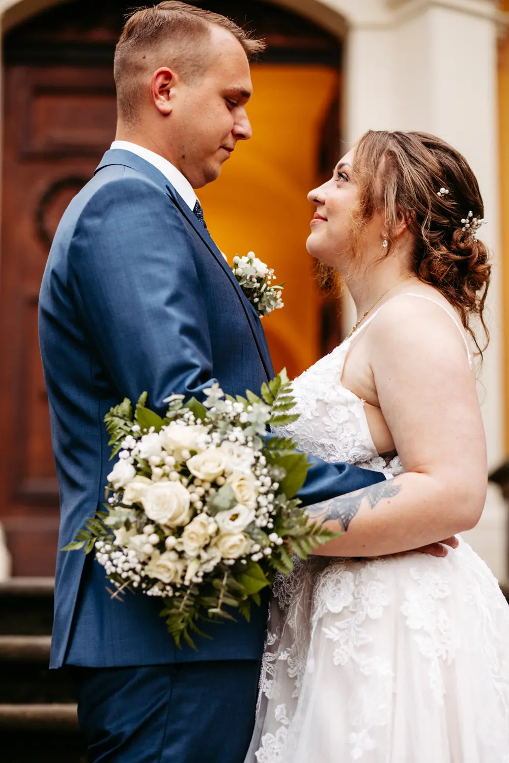 Eine Braut und ein Bräutigam stehen sich gegenüber und umarmen sich liebevoll. Der Bräutigam trägt einen blauen Anzug, die Braut ein weißes Spitzenkleid und hält einen Strauß aus weißen Rosen und Grünzeug in der Hand. Sie blicken sich vor einem Gebäude in die Augen.
