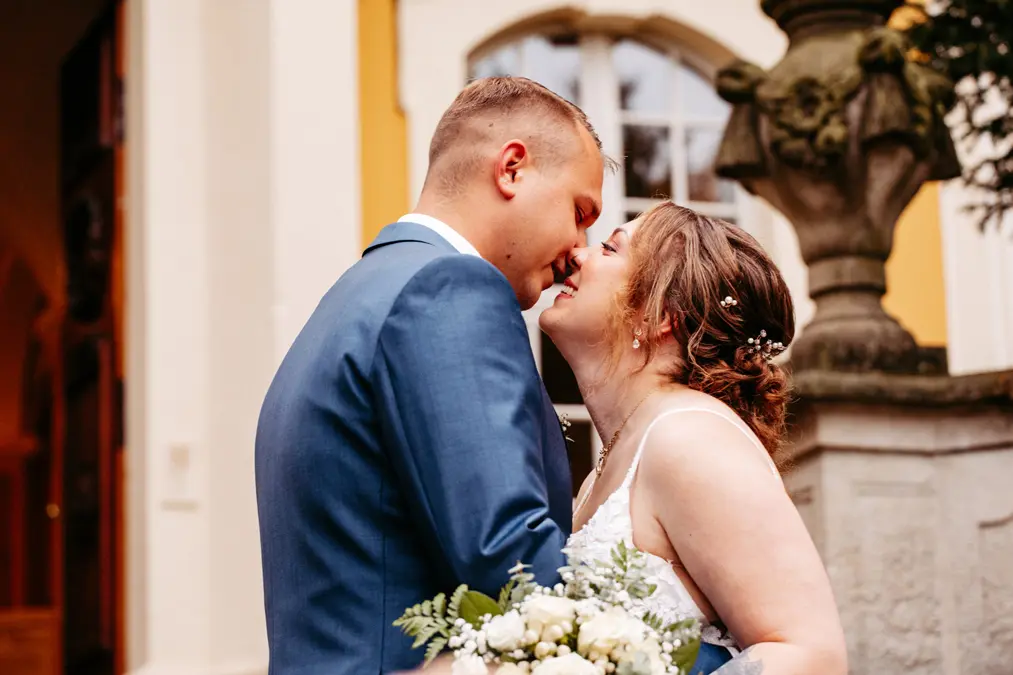 Eine Braut und ein Bräutigam lächeln sich liebevoll an, während sie draußen dicht beieinander stehen. Die Braut hält einen Strauß weißer Blumen und Grünzeug in der Hand, beide tragen formelle Hochzeitskleidung.