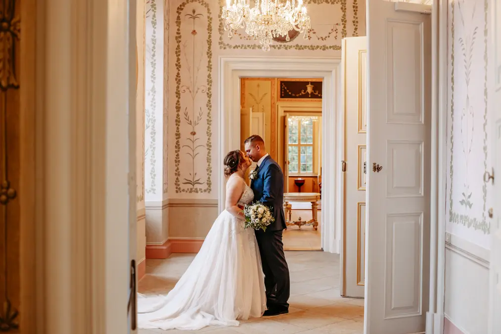 Eine Braut und ein Bräutigam stehen dicht beieinander in einem eleganten, kunstvoll dekorierten Flur und teilen einen intimen Moment. Die Braut trägt ein weißes Kleid und hält einen Blumenstrauß in der Hand, der Bräutigam trägt einen Anzug. Licht strömt durch große Fenster herein.