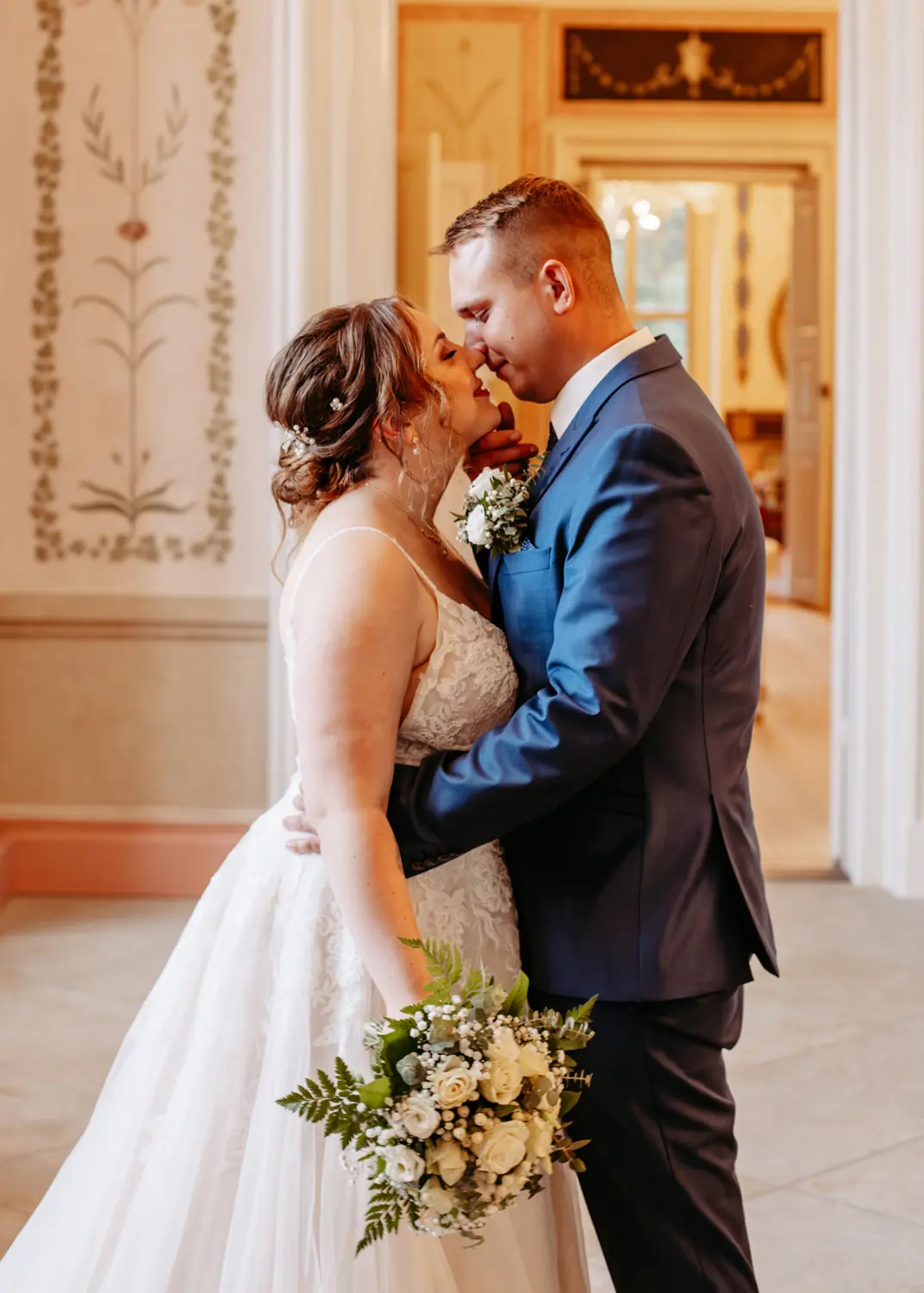 Eine Braut und ein Bräutigam stehen dicht beieinander, lächelnd und im Begriff, sich zu küssen. Die Braut trägt ein weißes Kleid und hält einen Strauß weißer Blumen, der Bräutigam trägt einen blauen Anzug. Sie befinden sich in einem Innenraum, mit eleganter Dekoration im Hintergrund.