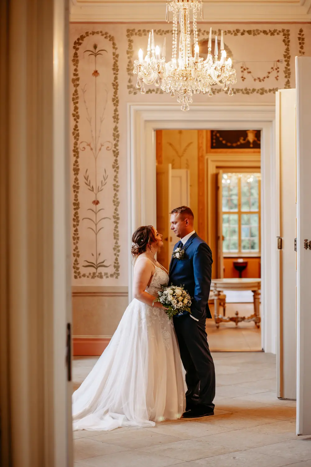 Eine Braut und ein Bräutigam stehen sich lächelnd in einem eleganten Raum mit verschnörkelten Wänden und einem Kronleuchter gegenüber. Die Braut trägt ein weißes Kleid und hält einen Blumenstrauß in der Hand, während der Bräutigam einen dunklen Anzug trägt.
