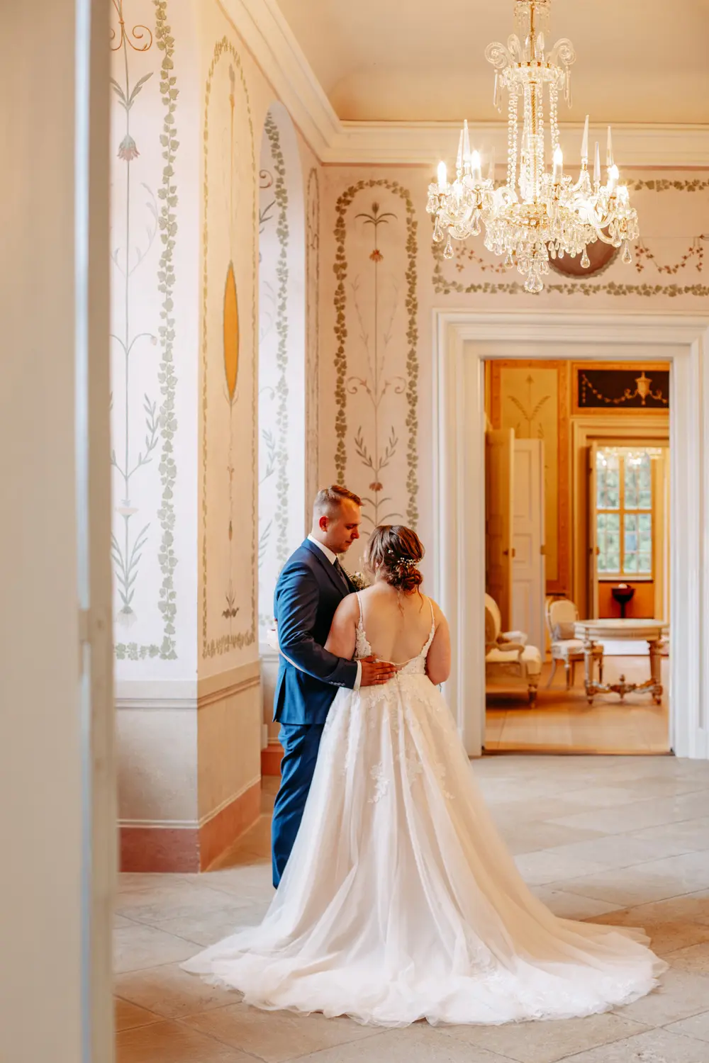 Eine Braut in einem weißen Kleid und ein Bräutigam in einem blauen Anzug stehen zusammen in einem eleganten, kunstvoll dekorierten Raum mit Kronleuchtern und floralen Wandgestaltungen und blicken auf einen angrenzenden, sonnenbeschienenen Raum.
