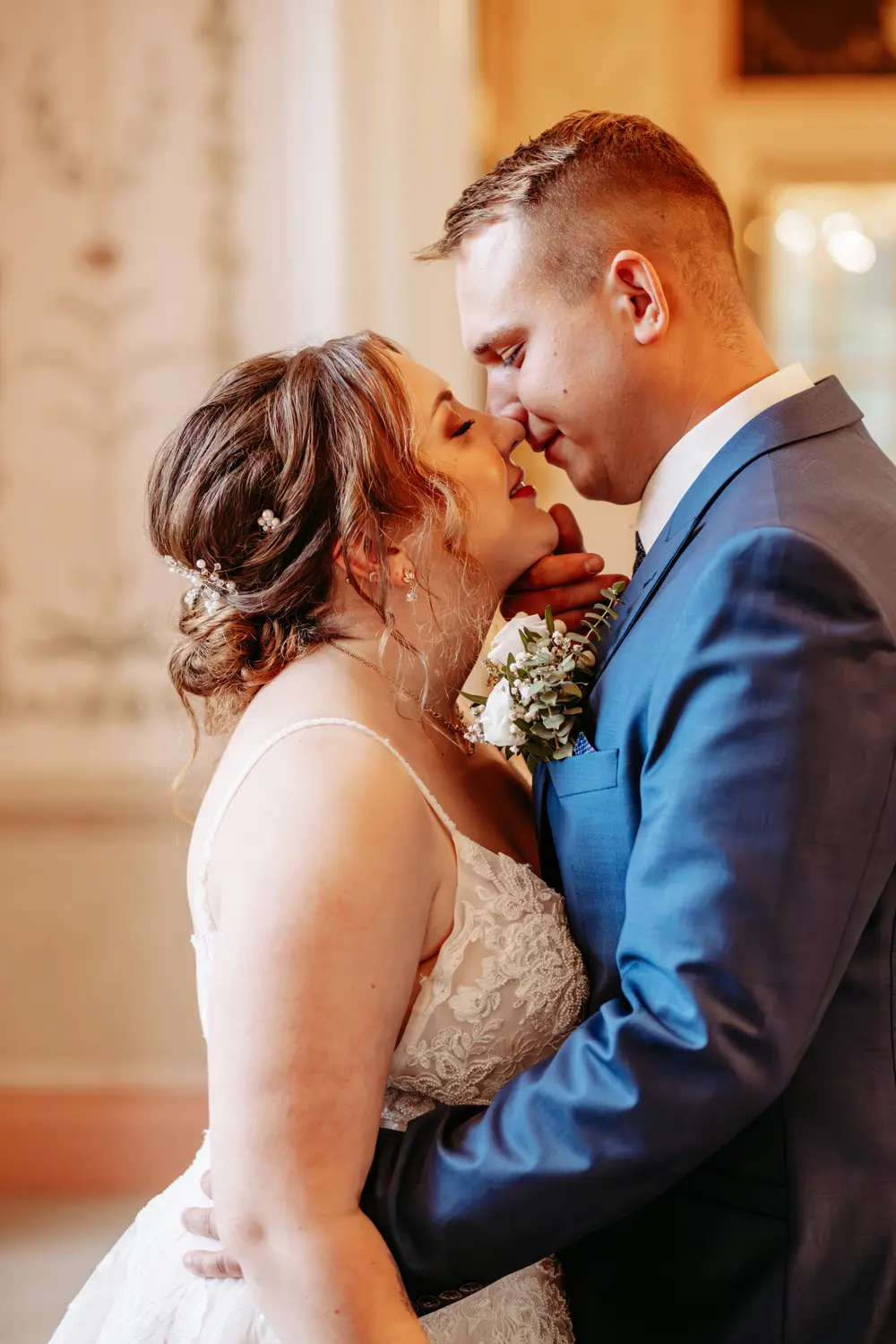 Eine Braut und ein Bräutigam stehen dicht beieinander, lächeln und berühren sich fast die Nasen, in einem zärtlichen Moment an ihrem Hochzeitstag. Die Braut trägt ein Spitzenkleid und der Bräutigam einen blauen Anzug mit einer Ansteckblume.