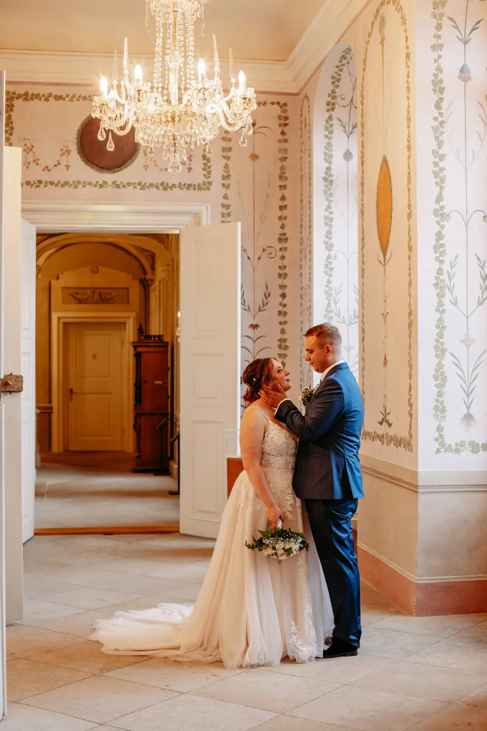 Eine Braut und ein Bräutigam stehen eng beieinander in einem elegant dekorierten Raum mit floralen Wandgestaltungen und einem Kronleuchter und schauen sich liebevoll an. Die Braut hält einen Blumenstrauß in der Hand.
