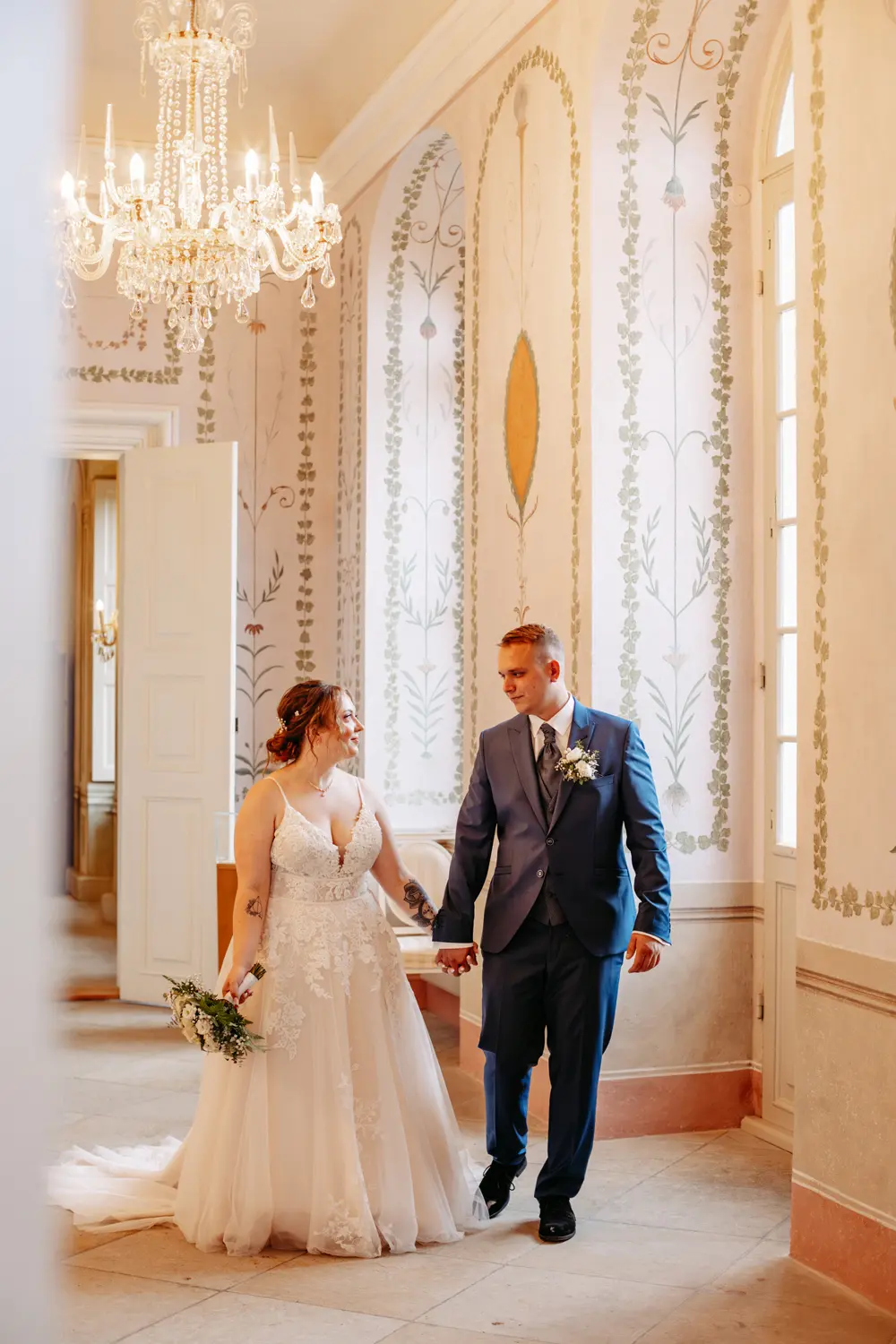 Eine Braut und ein Bräutigam halten sich an den Händen und lächeln sich an, während sie in einem Haus spazieren gehen. Die Braut trägt ein weißes Kleid und hat einen Blumenstrauß in der Hand, der Bräutigam trägt einen blauen Anzug. Der elegante Raum verfügt über hohe Fenster, verzierte Wände und einen Kronleuchter.