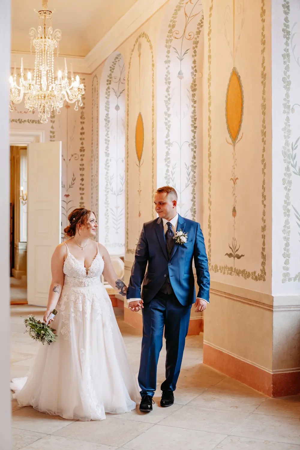 Eine Braut in einem weißen Spitzenkleid und ein Bräutigam in einem blauen Anzug halten sich an den Händen und gehen gemeinsam durch einen eleganten, kunstvoll verzierten Flur mit Kronleuchtern und detaillierten Wandgestaltungen.