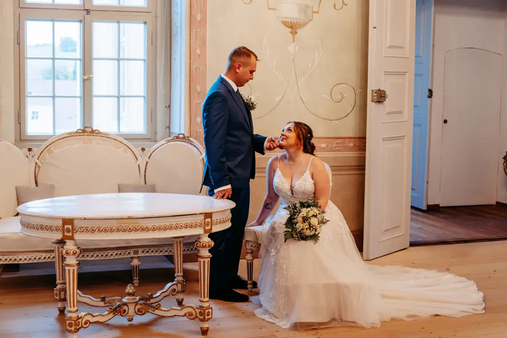 Eine Braut in einem weißen Kleid kniet mit einem Blumenstrauß in der Hand und lächelt zu einem Bräutigam in einem blauen Anzug auf, der sanft ihr Kinn berührt. Sie befinden sich in einem eleganten Raum mit verzierten Möbeln und hellen, pastellfarbenen Wänden.
