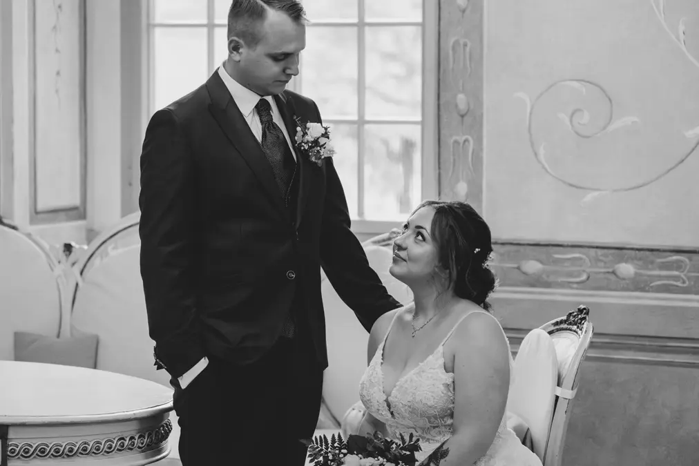 Ein Bräutigam im Anzug steht neben einer sitzenden Braut in einem weißen Kleid, die einen Blumenstrauß hält. Sie blicken sich liebevoll in einem eleganten Raum mit dekorativen Wänden und einem Fenster an. Das Foto ist in schwarz-weiß gehalten.