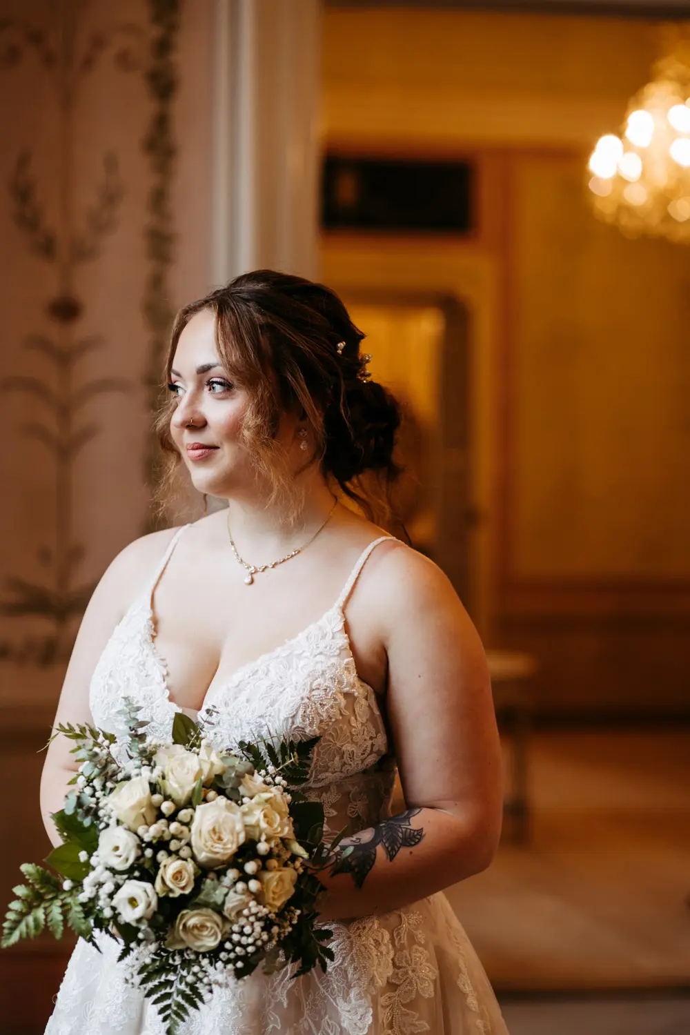 Eine Braut in einem weißen Spitzenhochzeitskleid, die einen Strauß weißer Rosen und Grünzeug hält, steht in einem Raum und blickt zur Seite. Im Hintergrund sind warme Beleuchtung und elegante Dekoration zu sehen.